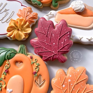 May include: An assortment of decorated fall-themed sugar cookies. The cookies include a red maple leaf with white speckles, an orange pumpkin, and a slice of pumpkin pie with whipped cream. Other cookies include a flower and a leaf.