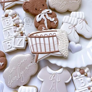 May include: A set of baby shower cookies decorated with a brown teddy bear, a crib, a baby onesie, and blocks that spell out "BABY". The cookies are decorated with white icing and have a light brown and white color scheme. The cookies are on a white surface and have the text "We can barely wait" on two of the cookies.