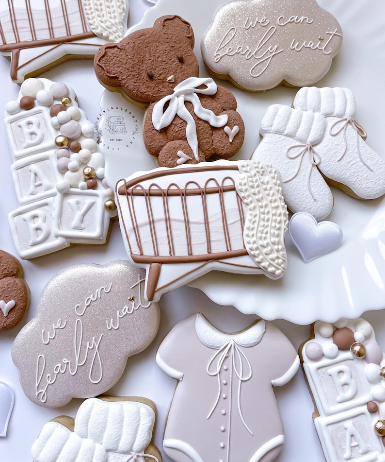 May include: A set of baby shower cookies decorated with a brown teddy bear, a crib, a baby onesie, and blocks that spell out "BABY". The cookies are decorated with white icing and have a light brown and white color scheme. The cookies are on a white surface and have the text "We can barely wait" on two of the cookies.