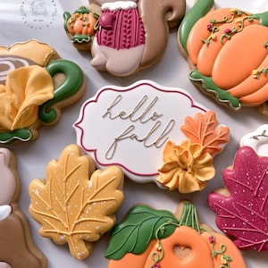 May include: An assortment of fall-themed cookies, including pumpkins, leaves, and a squirrel, decorated with orange, yellow, and red icing. A white cookie with the words "hello fall" in gold script is also present. The cookies are arranged on a white surface.