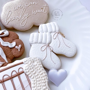 May include: A set of decorated sugar cookies with a white background. The cookies are decorated with white frosting and pink accents. One cookie is shaped like a cloud and says "we can barely wait". Another cookie is shaped like a pair of booties. A third cookie is shaped like a heart.