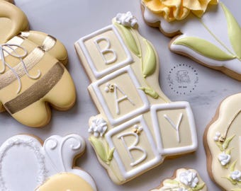 May include: Assortment of baby-themed cookies, including blocks spelling "BABY", a bee, and a flower. The cookies are decorated with white icing and yellow accents. The cookies are on a white surface.