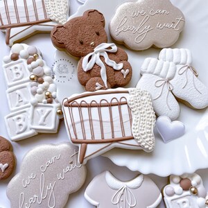 May include: A set of decorated sugar cookies for a baby shower. The cookies feature a brown teddy bear, a crib, a cloud with the text "we can barely wait", a baby onesie, and blocks that spell out "BABY".
