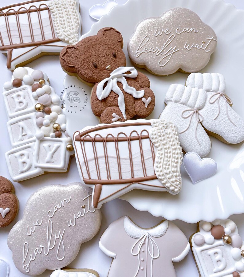 May include: A set of decorated sugar cookies for a baby shower. The cookies feature a brown teddy bear, a crib, a cloud with the text "we can barely wait", a baby onesie, and blocks that spell out "BABY".