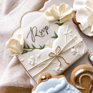 May include: A white cookie decorated with a gold border and white icing. The cookie is shaped like an envelope and has the word "RSVP" written on it in black cursive. The cookie is decorated with white flowers and green leaves.