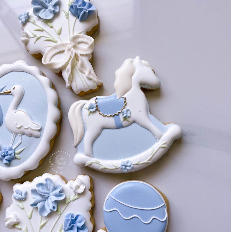 May include: Assortment of decorated sugar cookies in a light blue and white color scheme. Designs include a rocking horse, a stork, a bouquet of flowers, and a hot air balloon. The cookies are decorated with intricate details and floral accents.