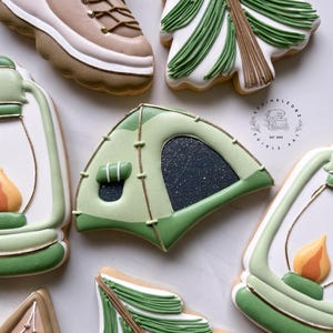 May include: Assortment of camping-themed cookies, including a tent, hiking boot, and lantern, decorated with green, white, and brown icing. The cookies have gold accents and are arranged on a white surface. The cookies are decorated with intricate details.