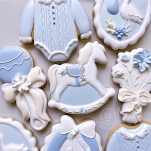 May include: An assortment of baby-themed cookies, including a onesie, a rocking horse, a rattle, and a bouquet of flowers. The cookies are decorated with light blue and white icing, with intricate details and designs. The cookies are arranged on a light-colored surface.