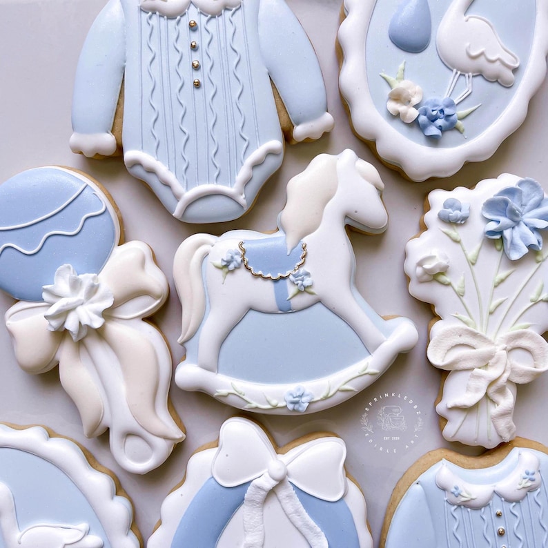 May include: An assortment of baby-themed cookies, including a onesie, a rocking horse, a rattle, and a bouquet of flowers. The cookies are decorated with light blue and white icing, with intricate details and designs. The cookies are arranged on a light-colored surface.