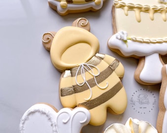 May include: A collection of decorated sugar cookies. One cookie is shaped like a bee with yellow and gold stripes. Other cookies include a cake, a dress, and a flower. The cookies are decorated with white and gold icing.
