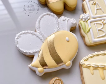 May include: Close-up of decorated sugar cookies. A yellow bee-shaped cookie with gold stripes and white wings is in the center. Other cookies include letter blocks and a bear. The cookies are decorated with white and gold icing.