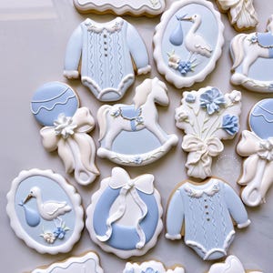 May include: An assortment of baby-themed cookies, decorated with light blue and white icing. Designs include a onesie, a stork carrying a baby, a rocking horse, a rattle, and a floral bouquet. The cookies are arranged on a white surface.