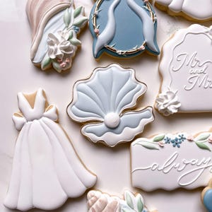 May include: A collection of decorated sugar cookies featuring a white wedding dress, a blue seashell with a pearl, a plaque with the text Mr and Mrs, a plaque with the text always, and floral accents in shades of blue, pink, and green with gold trim.