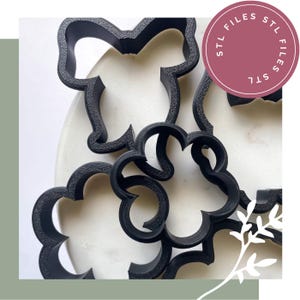 May include: A collection of black plastic cookie cutters with various shapes, including a flower and a bow design. A circular pink badge in the upper right corner features the repeated white text STL FILES STL FILES STL.