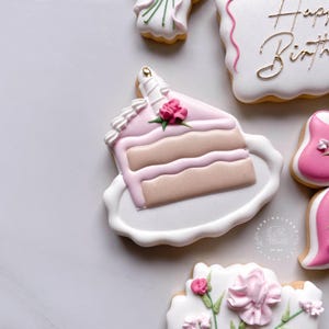 May include: Decorated sugar cookies with a birthday theme. A slice of cake cookie with pink frosting and a rose sits on a white plate. Other cookies include a floral design and a cookie with "Happy Birthday" in gold lettering.