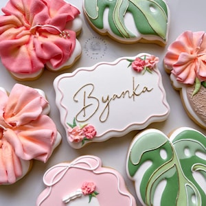May include: Decorated sugar cookies with a floral theme. The cookies include pink flowers, green monstera leaves, and a cookie with the name "Byanka" in gold script. The cookies are decorated with white, pink, and green icing.