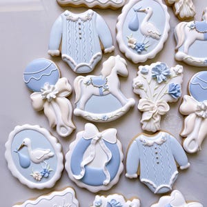 K&ouml;nnte beinhalten: Eine Auswahl an Baby-Themen-Keksen in Hellblau und Wei&szlig;. Designs umfassen einen Baby-Strampler, einen Storch mit einem B&uuml;ndel, ein Schaukelpferd, eine Rassel und Blumenarrangements. Die Kekse sind mit aufwendigen Details verziert.