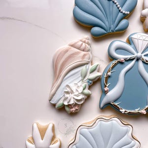 May include: A collection of decorated sugar cookies featuring a large beige conch shell with white floral icing, a blue shell, a blue ring-shaped cookie with a white diamond and bow, and a white shell cookie, all with intricate piped icing details.