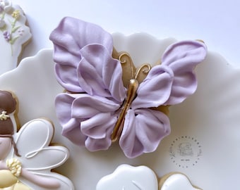May include: A decorated butterfly cookie with lavender-colored wings and gold accents. The cookie is on a white plate with other decorated cookies, including a fairy and flower-shaped cookies. The cookie is part of a set of edible art.