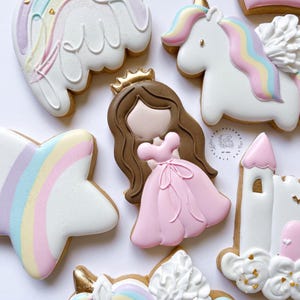May include: A set of decorated sugar cookies featuring a princess, a unicorn, a rainbow, a star, and a castle. The cookies are decorated with pink, blue, white, and gold icing.