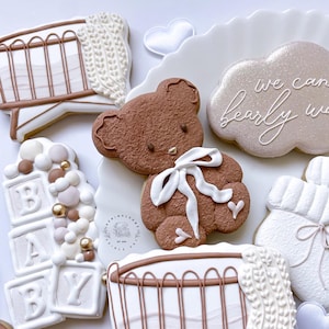 May include: A set of six decorated sugar cookies featuring a brown teddy bear, a crib, a cloud with the text "We can barely wait", a stack of blocks spelling "BABY", and two other cookies with a knitted texture.