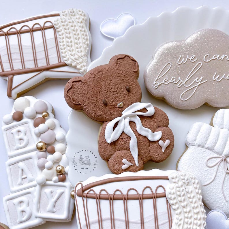 May include: A set of six decorated sugar cookies featuring a brown teddy bear, a crib, a cloud with the text "We can barely wait", a stack of blocks spelling "BABY", and two other cookies with a knitted texture.
