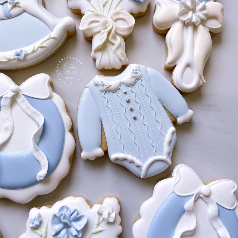 K&ouml;nnte beinhalten: Eine Auswahl an Baby-Themen-Keksen. Die Kekse sind mit hellblauer und wei&szlig;er Glasur verziert und zeigen Designs wie einen Strampler, ein Schaukelpferd, Schleifen und florale Akzente. Die Kekse sind auf einer hellen Oberfl&auml;che angeordnet.