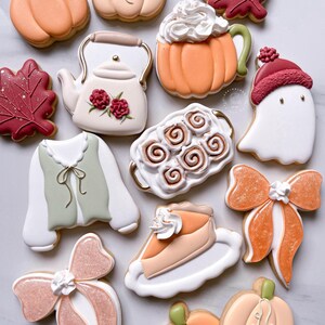 May include: An assortment of decorated sugar cookies in fall and autumn themes. Cookies include pumpkins, fall leaves, a teapot, a ghost, a slice of pie, bows, and a mug of pumpkin spice latte.