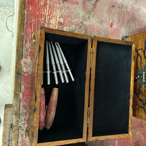 May include: A wooden box with a black velvet interior, containing a set of silver crochet hooks and a pink and brown crochet hook. The box has a brass latch and hinges.