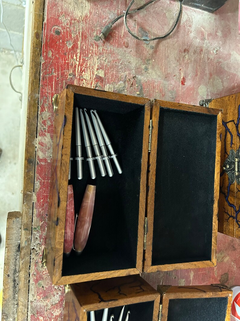May include: A wooden box with a black velvet interior, containing a set of silver crochet hooks and a pink and brown crochet hook. The box has a brass latch and hinges.