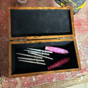May include: A wooden box with a black velvet interior, containing a set of silver crochet hooks and two pink and purple crochet hooks. The box is open and the hooks are visible.