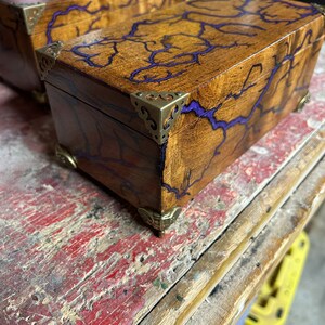 May include: Two wooden boxes with purple resin inlay and brass corner accents. The boxes are on a wooden surface.