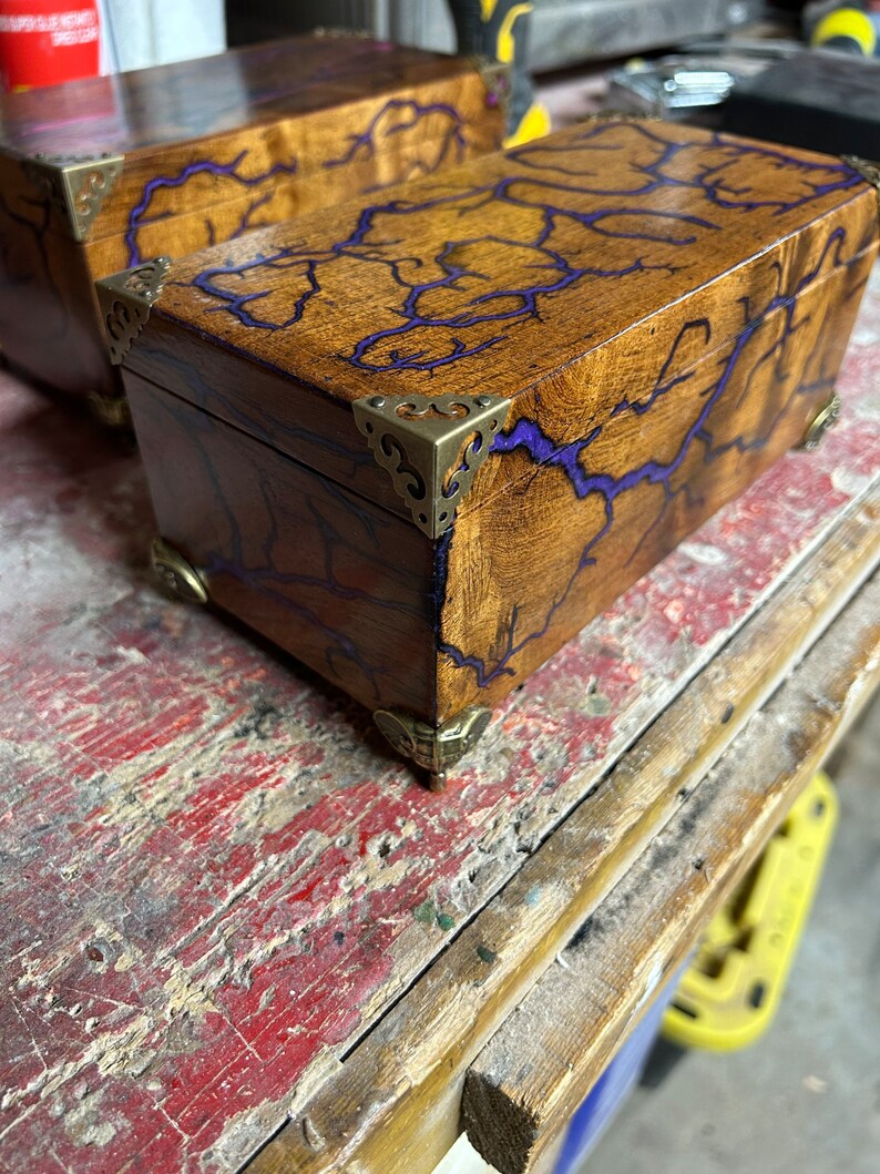 May include: Two wooden boxes with purple resin inlay and brass corner accents. The boxes are on a wooden surface.