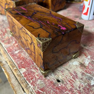 May include: A wooden box with a purple and pink resin inlay. The box has brass corner accents and a decorative brass hinge.