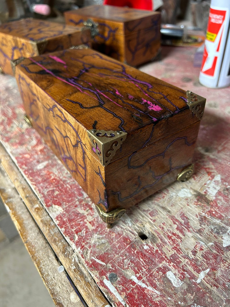 May include: A wooden box with a purple and pink resin inlay. The box has brass corner accents and a decorative brass hinge.
