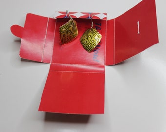 May include: A pair of gold earrings with a geometric design and black accents. The earrings are in a red box with a white and blue pattern.