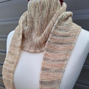 Könnte beinhalten: Handgestrickter Schal in Beige-, Pfirsich- und Brauntönen. Der Schal mit strukturiertem Strickmuster wird auf einer Schaufensterpuppe präsentiert. Die Enden sind lang und spitz zulaufend. Eine bordeauxfarbene Blume schmückt den Kopf der Puppe.