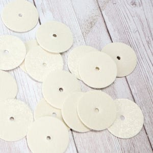 May include: A pile of off-white felt polishing pads with a hole in the center. The pads are arranged in a random pattern on a light wood surface.
