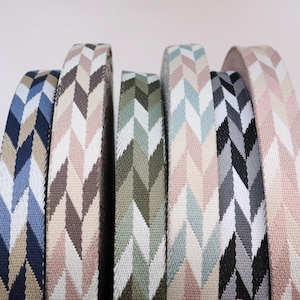 May include: Close-up of several rolls of patterned fabric ribbon. The ribbons feature a chevron design in various colors, including navy blue, light blue, tan, pink, olive green, black, and gray. The ribbons are tightly rolled.