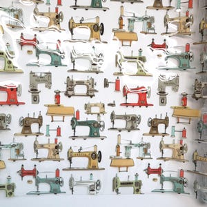 May include: A white fabric featuring a repeating pattern of vintage sewing machines in various colors including red, teal, yellow, and grey, interspersed with small red and teal thread spools arranged in a grid-like layout across the material.