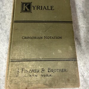 May include: A vintage olive green hardcover book titled Kyriale Gregorian Notation. The cover features black text, a decorative letter K, and the publisher name J. Fischer & Brother New York printed in black ink with horizontal line accents.
