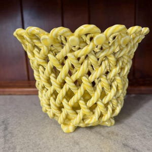 May include: A yellow ceramic basket with a woven lattice design. The bowl features a flared rim and a base supported by small feet. The surface has a glossy finish with white highlights along the twisted rope-like ceramic strands.