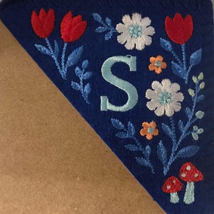 May include: A blue felt pennant with embroidered floral and mushroom designs. The pennant features red tulips, white and orange flowers, and a large embroidered letter "S" in light blue. The edges are stitched with a decorative border.