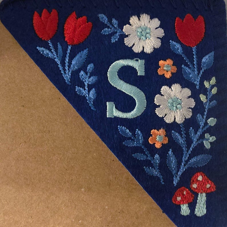 May include: A blue felt pennant with embroidered floral and mushroom designs. The pennant features red tulips, white and orange flowers, and a large embroidered letter "S" in light blue. The edges are stitched with a decorative border.
