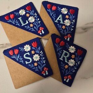 May include: Four navy blue felt bookmarks with embroidered floral designs and letters. Each bookmark features red tulips, white flowers, and small orange blossoms. The letters are in light blue. The bookmarks are on brown paper.