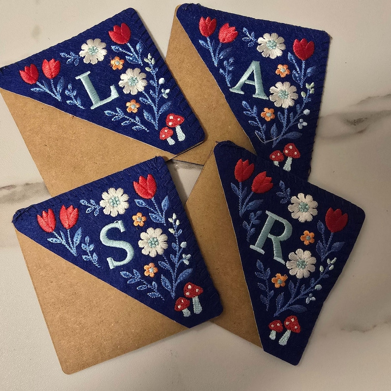 May include: Four navy blue felt bookmarks with embroidered floral designs and letters. Each bookmark features red tulips, white flowers, and small orange blossoms. The letters are in light blue. The bookmarks are on brown paper.