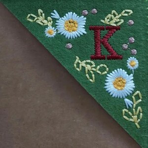 May include: Green felt bookmark with embroidered floral and leaf designs. The bookmark features a large, embroidered red letter "K" and white daisies with yellow centers. The edges are trimmed with embroidered leaves and small pink dots.