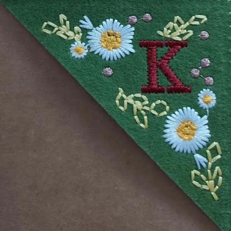 May include: Green felt bookmark with embroidered floral and leaf designs. The bookmark features a large, embroidered red letter "K" and white daisies with yellow centers. The edges are trimmed with embroidered leaves and small pink dots.