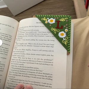 May include: Green felt bookmark with embroidered white daisies, yellow centers, and the letter "L". The bookmark is placed in a book, with the text "THE SPAN" visible.