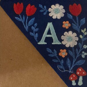 May include: A blue felt banner with embroidered floral and mushroom designs. The banner features the letter "A" in light blue, surrounded by red tulips, white and orange flowers, and blue leaves. Two red mushrooms with white spots are at the bottom.